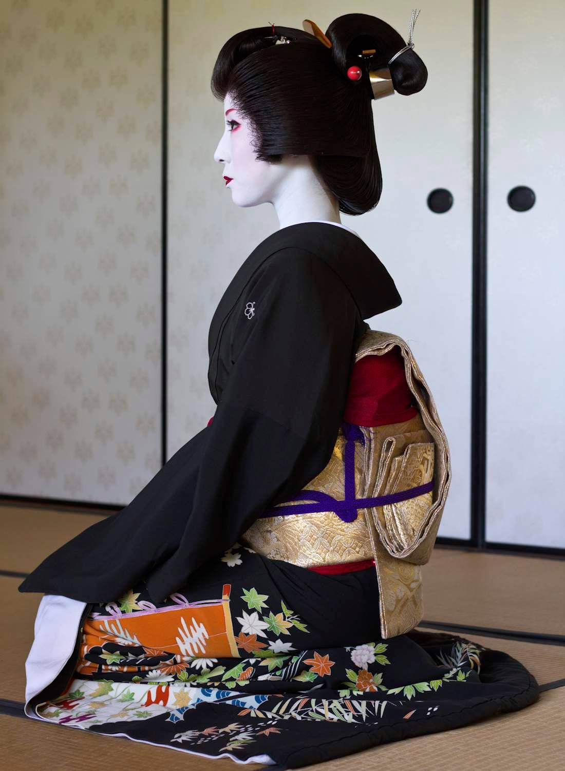 Portrait de profil d'une Geiko de Kyoto assise en position traditionnelle seiza, portant un kimono noir formel avec un obi doré et un maquillage blanc shironuri.