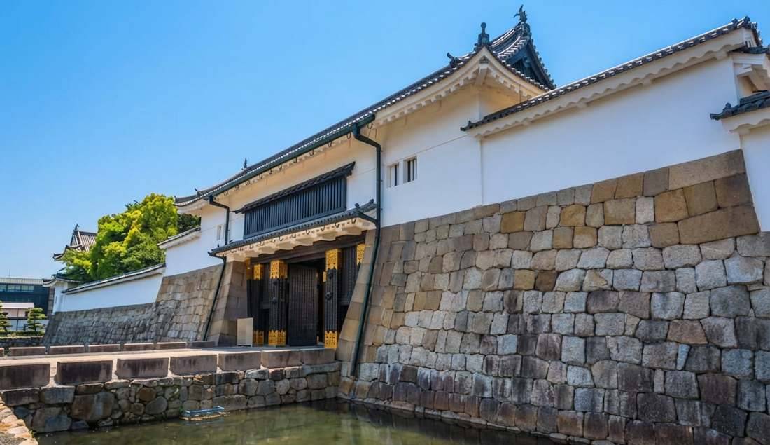 Porte principale Higashi Ote-mon du château de Nijo à Kyoto avec ses murs en pierre massifs, ses dorures traditionnelles et ses douves sous un ciel bleu clair.