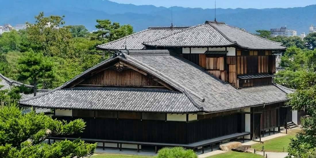 Vue extérieure du palais Honmaru au château de Nijo à Kyoto, Japon, montrant l'architecture traditionnelle en bois, les toits de tuiles grises et les jardins environnants sous un ciel bleu.