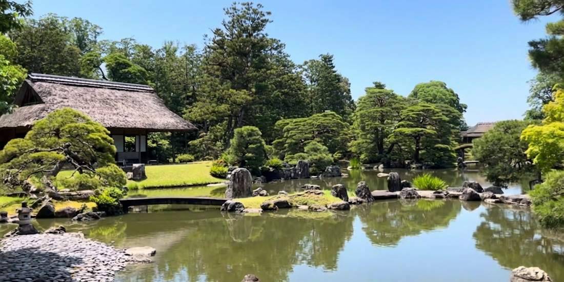 Vue panoramique du jardin japonais de la Villa Impériale Katsura à Kyoto avec son étang central, un pavillon de thé traditionnel au toit de chaume, un pont en pierre et des pins soigneusement taillés sous un ciel bleu.