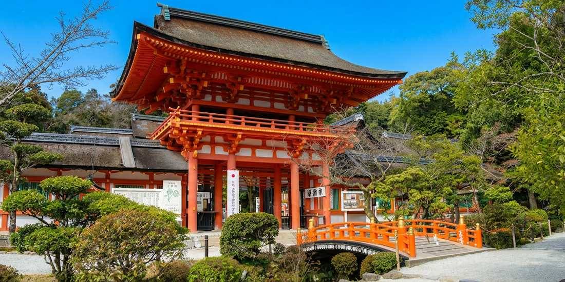 Vue de la porte monumentale vermillon (Romon) et du petit pont cintré du sanctuaire Kamigamo-jinja à Kyoto, entourés de pins et de jardins traditionnels japonais sous un ciel bleu.