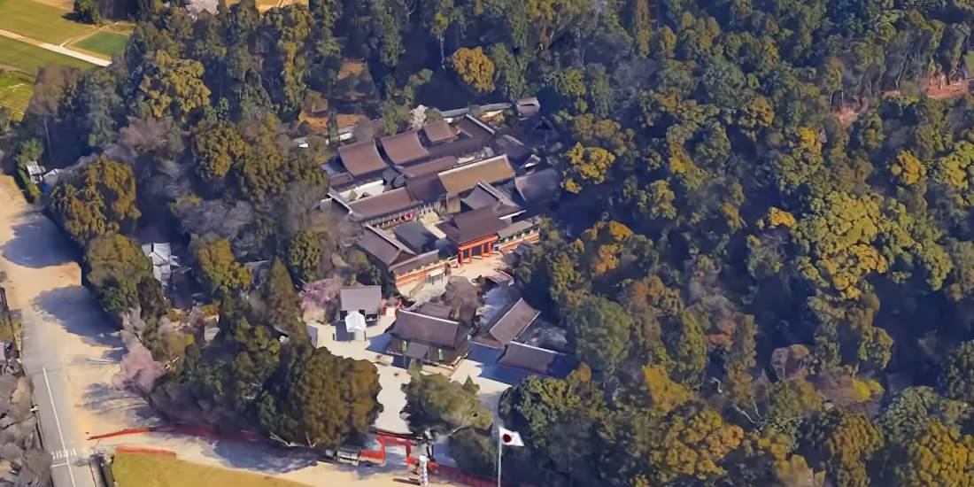 Cette photographie aérienne présente le complexe du sanctuaire Kamigamo-jinja (Kamo-wakeikazuchi), situé dans le nord de Kyoto. On y distingue clairement l'agencement complexe des bâtiments traditionnels aux toits de bardeaux de cyprès, les emblématiques portails torii vermillon et l'intégration harmonieuse de l'architecture dans le paysage naturel boisé. En tant que site classé au patrimoine mondial de l'UNESCO, cette image illustre parfaitement la sérénité et l'importance historique de la culture shintoïste au Japon. Vue aérienne du sanctuaire shinto Kamigamo-jinja entouré d'une forêt dense à Kyoto, Japon, montrant l'architecture traditionnelle des toits et les portes torii rouges.