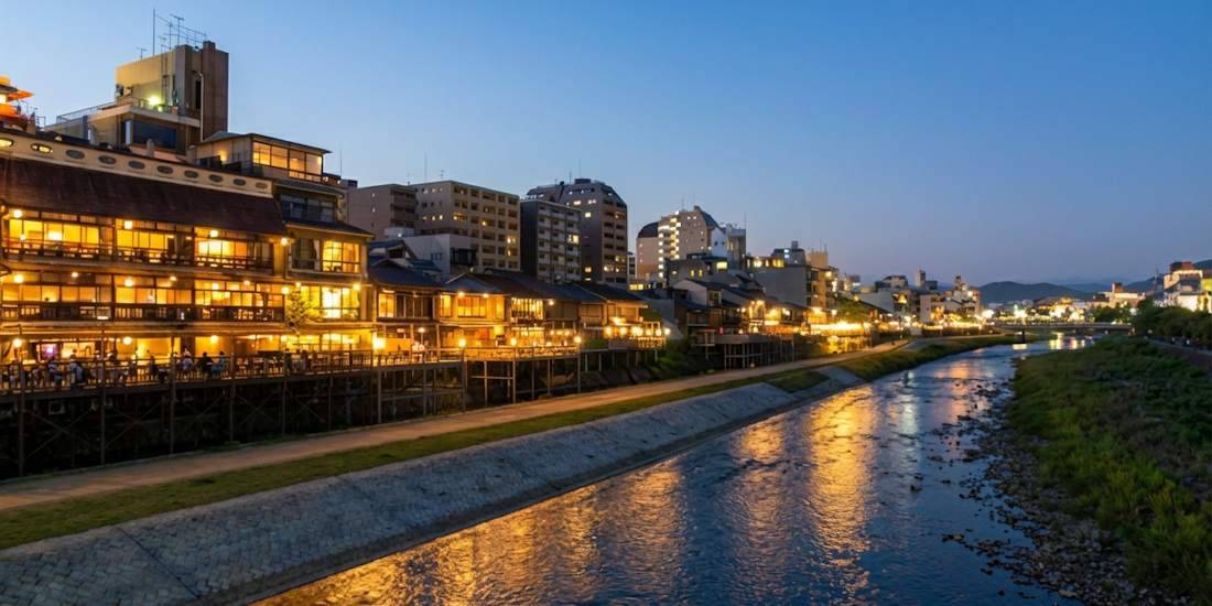 Vue nocturne des terrasses en bois (yuka) du quartier de Pontocho bordant la rivière Kamogawa à Kyoto, Japon, avec les lumières des restaurants se reflétant sur l'eau au crépuscule.