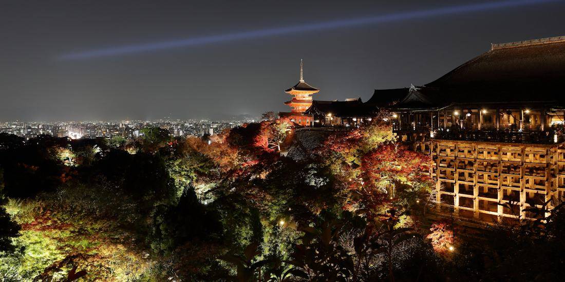 Vue nocturne du temple Kiyomizu-dera à Kyoto avec sa célèbre plateforme en bois, la pagode illuminée et les érables rouges d'automne sous un faisceau lumineux bleu.