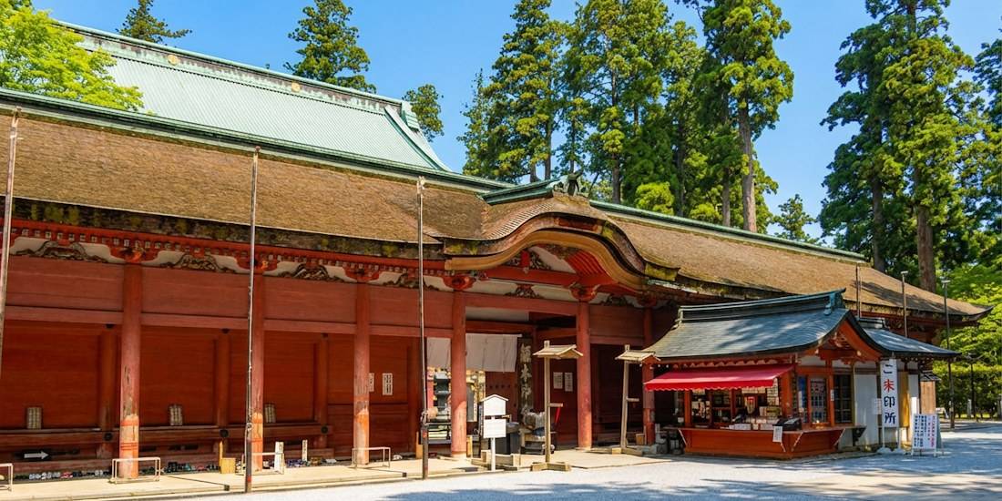 Façade en bois rouge du temple Konpon Chū-dō à l'Enryaku-ji sur le mont Hiei près de Kyoto, entourée de grands cèdres sous un ciel bleu.