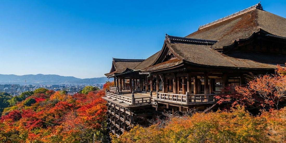 Temple Kiyomizu-dera à Kyoto entouré de feuilles d'automne rouges et orange avec vue panoramique sur la ville sous un ciel bleu.