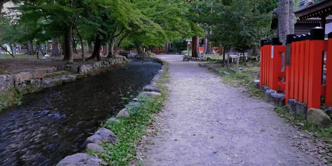 Cette photographie capture la sérénité du ruisseau Nara-no-Ogawa, situé dans le complexe du sanctuaire shinto Kamigamo-jinja à Kyoto. On y voit un sentier de terre longeant l'eau claire, entouré d'une végétation luxuriante et d'arbres centenaires. À droite, les clôtures traditionnelles d'un rouge vermillon éclatant contrastent avec les tons verts et gris du paysage, illustrant parfaitement l'esthétique spirituelle et naturelle du Japon traditionnel. Ce lieu est célèbre pour ses rituels de purification et son atmosphère paisible, classé au patrimoine mondial de l'UNESCO. Ruisseau Nara-no-Ogawa bordé d'arbres et d'une clôture vermillon au sanctuaire Kamigamo-jinja à Kyoto, Japon.