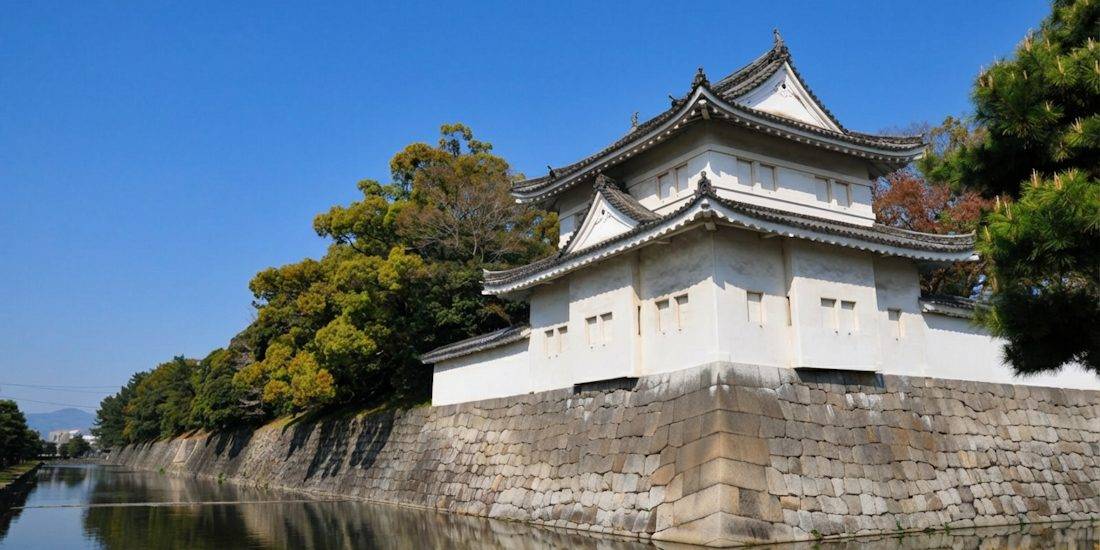 Vue extérieure du château de Nijo à Kyoto, Japon, montrant une tour de guet blanche (yagura) sur des murs en pierre massifs bordant les douves sous un ciel bleu clair.