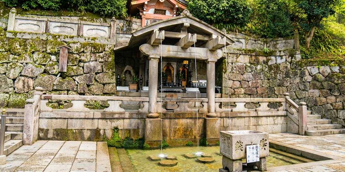 Cascade Otowa-no-taki au temple bouddhiste Kiyomizu-dera à Kyoto, Japon, montrant les trois canaux d'eau sacrée et l'architecture traditionnelle en pierre entourée de végétation.