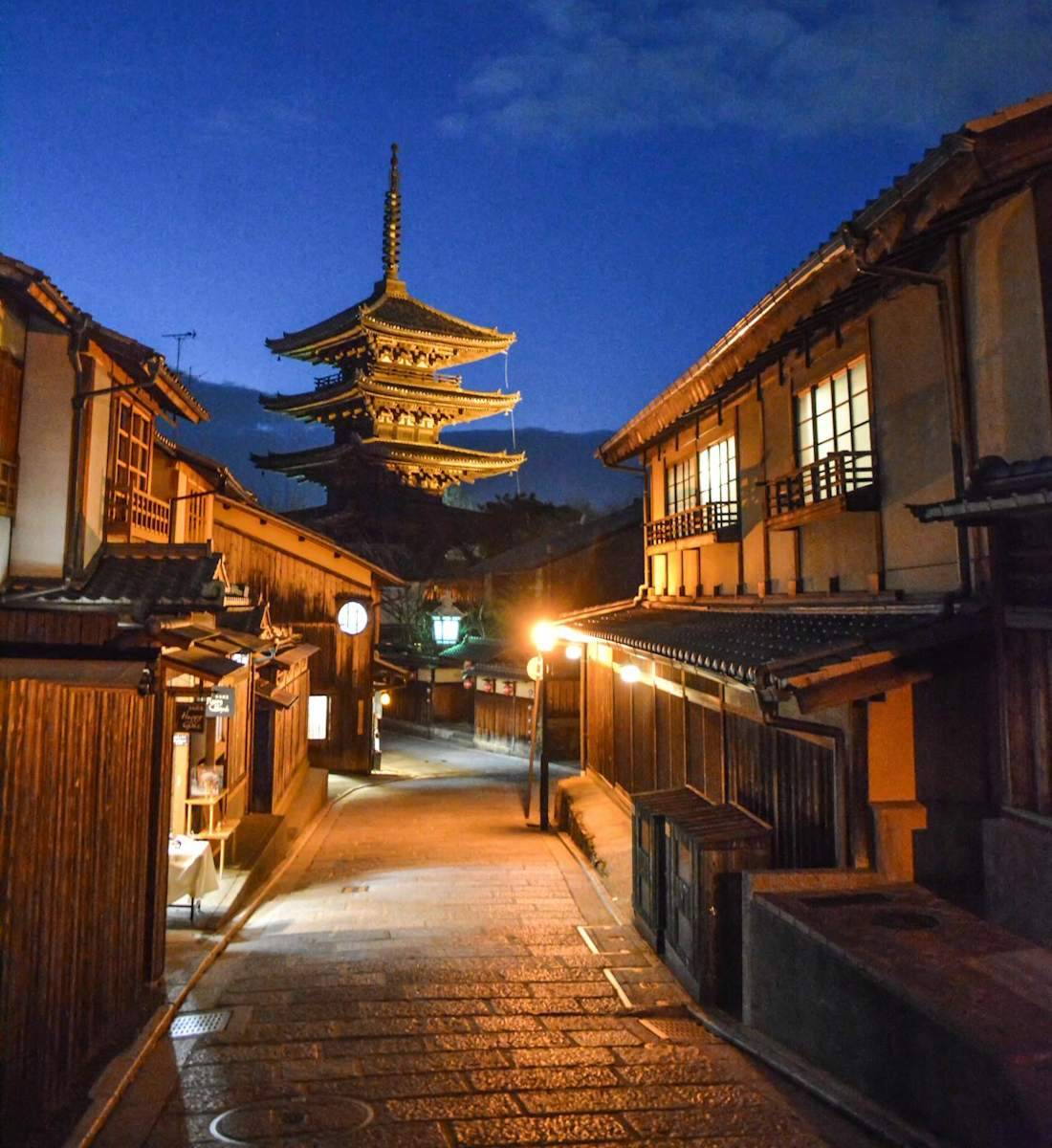 Une rue pavée traditionnelle du quartier de Gion à Kyoto de nuit, avec la pagode Yasaka-no-to illuminée en arrière-plan sous un ciel bleu profond.