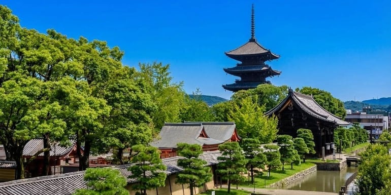 Pagode à cinq étages du temple To-ji à Kyoto, Japon, entourée de jardins verdoyants et de bâtiments traditionnels sous un ciel bleu clair.