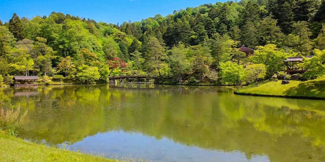 Pont en bois traditionnel traversant l'étang du jardin supérieur de la Villa Impériale Shugaku-in à Kyoto, entouré d'une forêt luxuriante sous un ciel bleu.