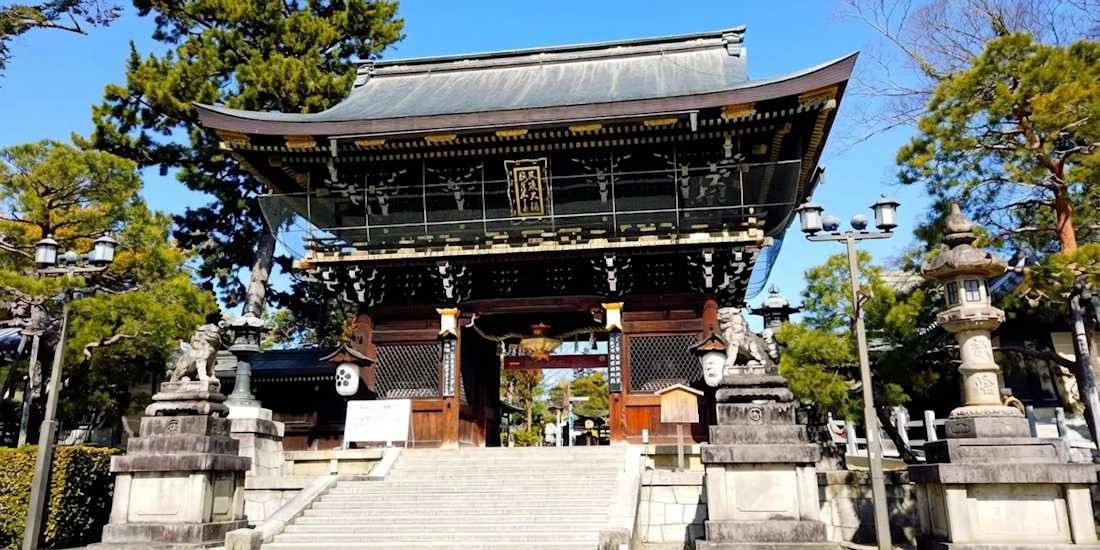 Porte Sankomon monumentale en bois sculpté à l'entrée du sanctuaire Kitano Tenman-gu à Kyoto, Japon, sous un ciel bleu avec des statues de lions gardiens Komainu au premier plan.