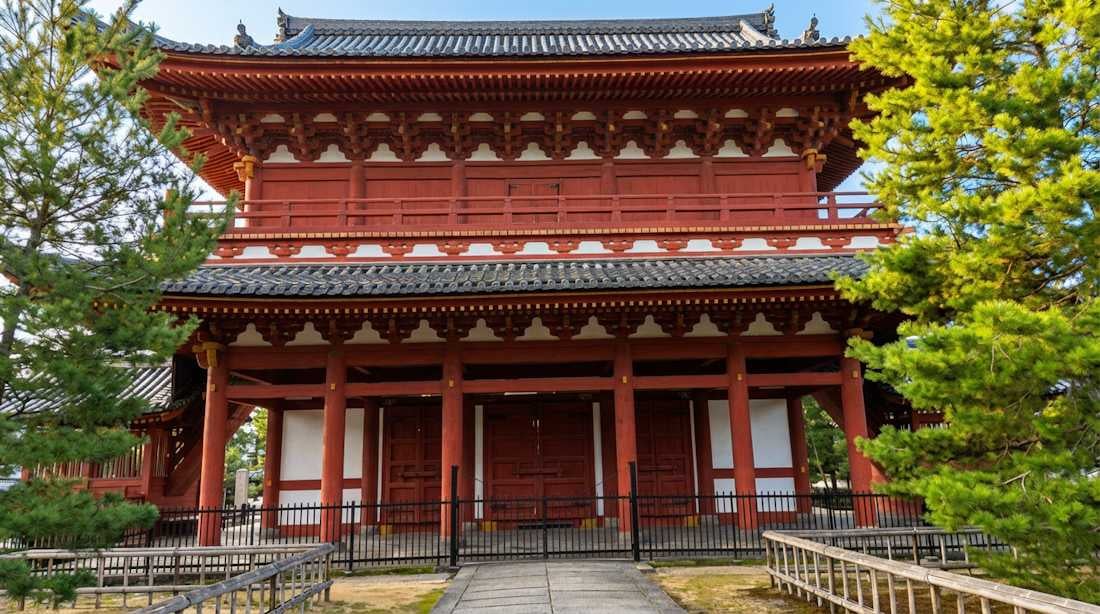 Porte Sanmon monumentale en bois rouge du temple Myoshin-ji à Kyoto, Japon, entourée de pins sous un ciel clair.