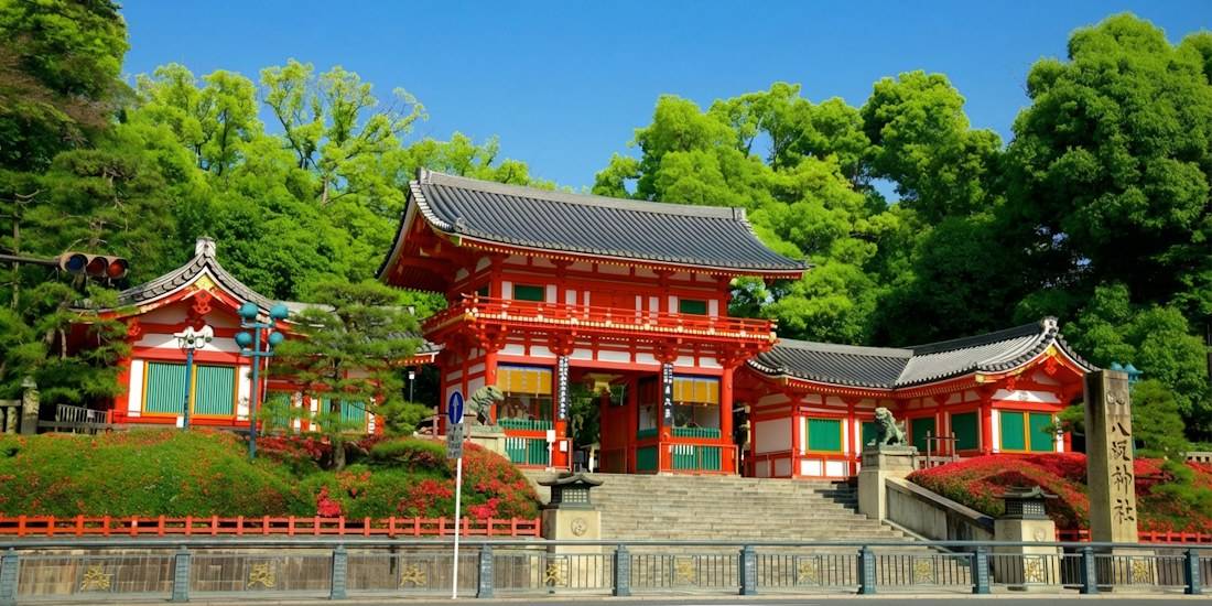 Vue de la porte principale vermillon Nishi-romon du sanctuaire Yasaka-jinja à Kyoto, Japon, entourée d'arbres verdoyants sous un ciel bleu clair.