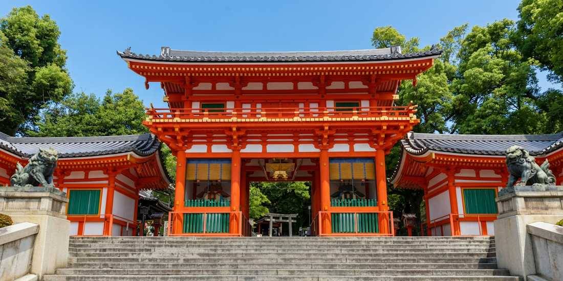 Entrée principale du sanctuaire Yasaka-jinja à Kyoto, montrant la porte vermillon Nishi-romon avec ses statues de lions gardiens komainu sous un ciel bleu.