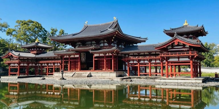 Vue panoramique du Pavillon du Phénix (Ho-o-do) du temple Byodo-in à Uji, Kyoto, avec son architecture rouge emblématique se reflétant sur l'eau sous un ciel bleu clair.