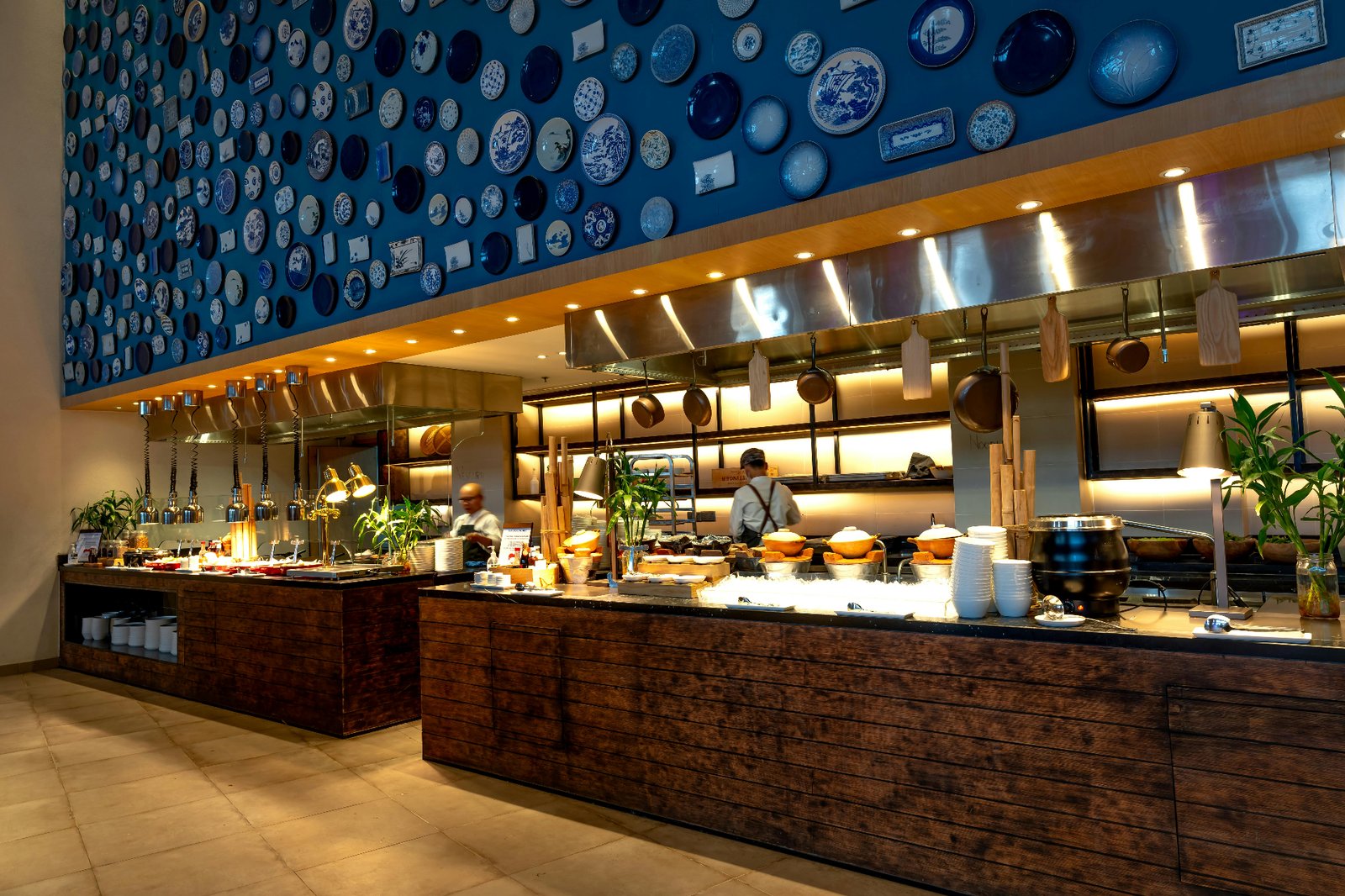 Open restaurant kitchen with chefs working an exhibition line under a decorative blue plate wall, reflecting modern restaurant operations and guest-facing service