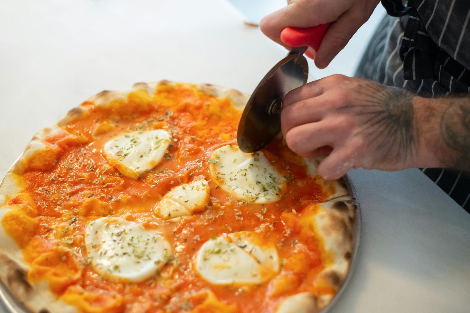 Pizzaiolo slicing a fresh margherita pizza with mozzarella and oregano using a wheel cutter