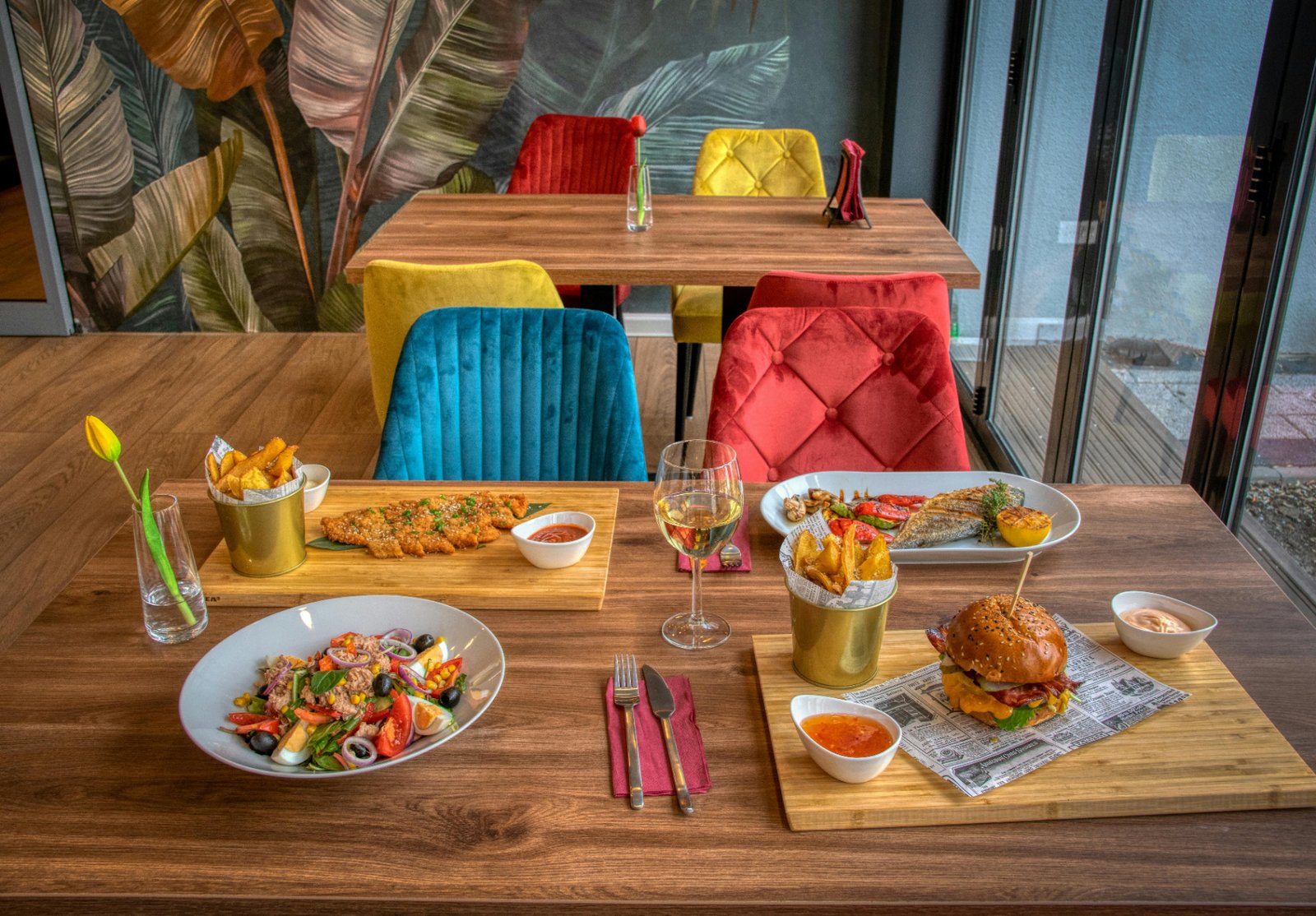 Bright restaurant table set with burger, fish, salad, and wine in front of colorful velvet chairs