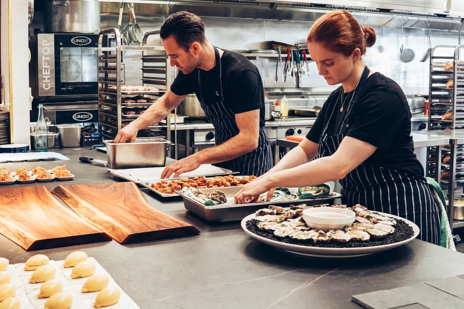 Two chefs in striped aprons plating oysters and pastries on a stainless steel kitchen pass, illustrating smarter back-of-house operations