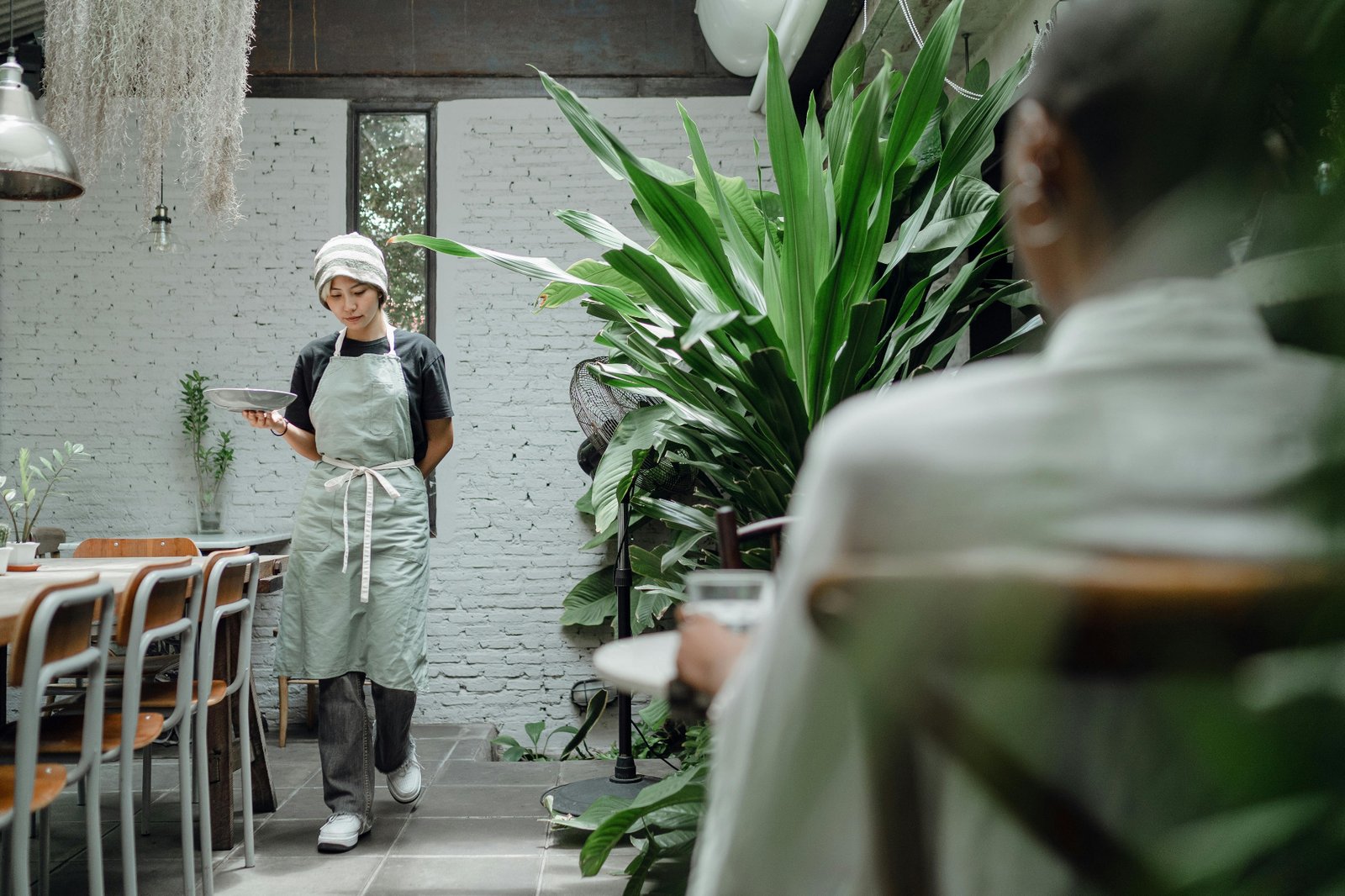 Restaurant server in apron carrying a clean plate through a dining room with plants and brick wall