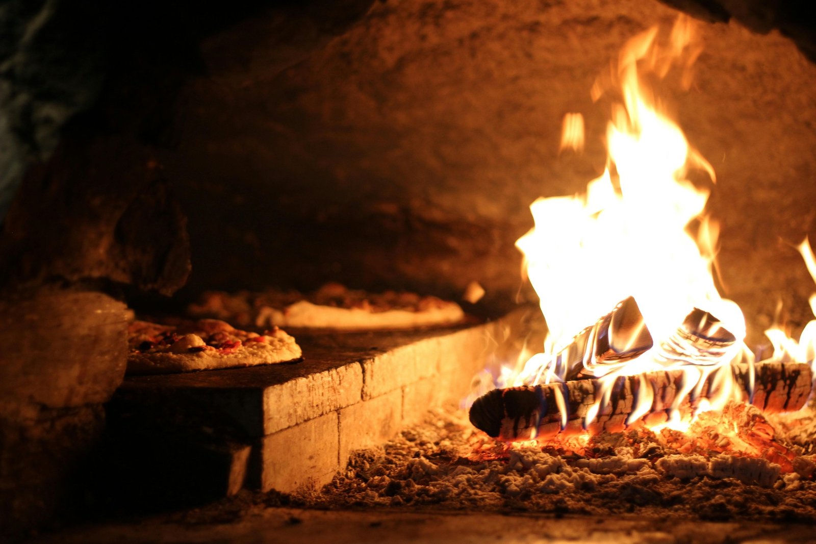 Pizzas baking on the stone floor of a traditional wood-fired pizza oven with roaring flames