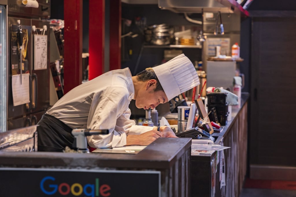 A chef in a white toque writing down orders at the counter of a busy restaurant kitchen.