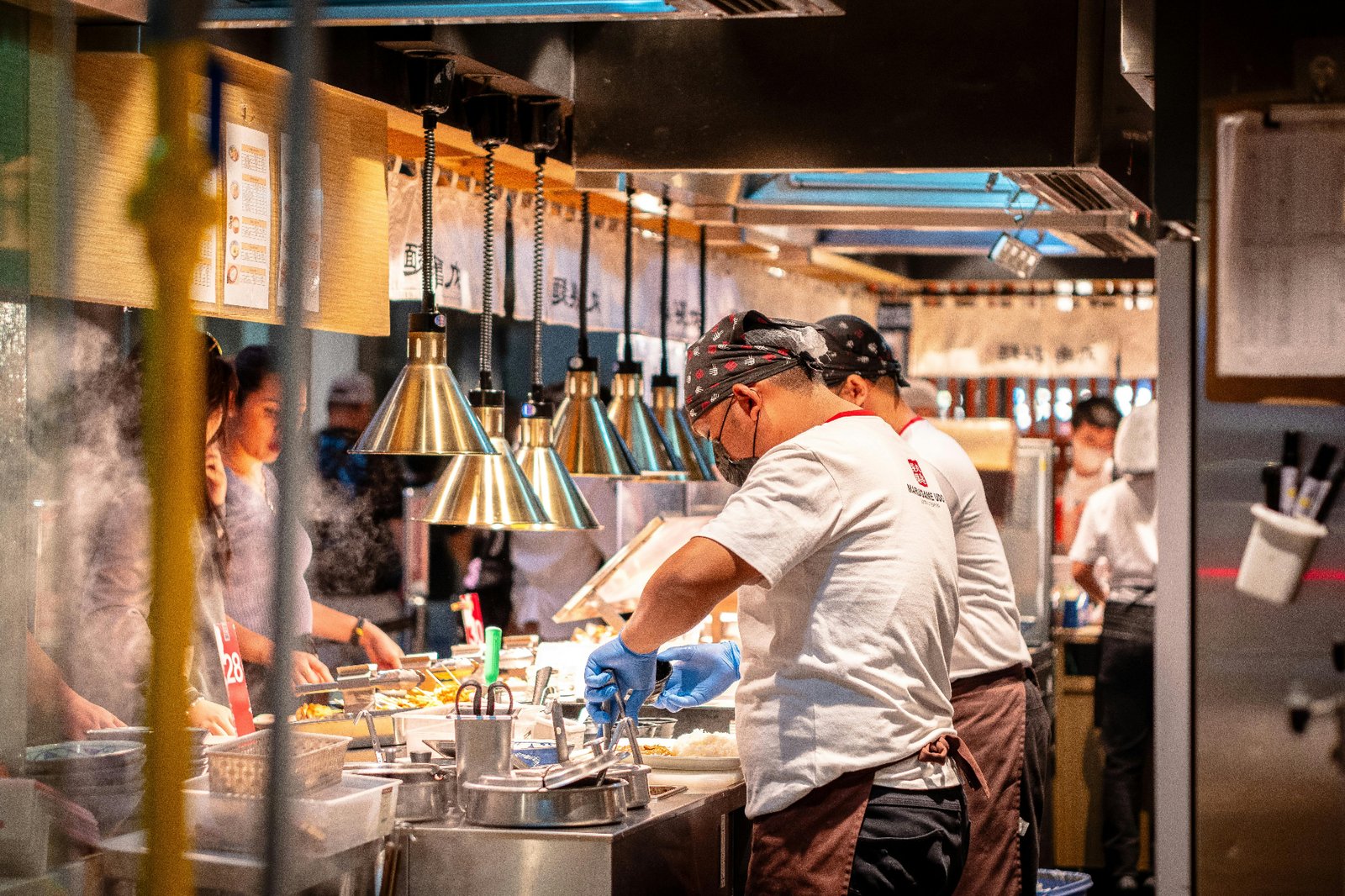 Line cook plating dishes under heat lamps in a busy open kitchen