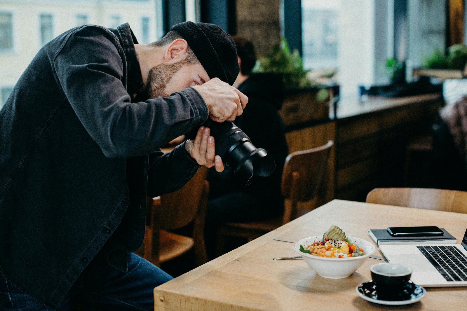 Restaurant food photographer shooting a styled bowl on a wooden cafe table next to a laptop and notebook for marketing content