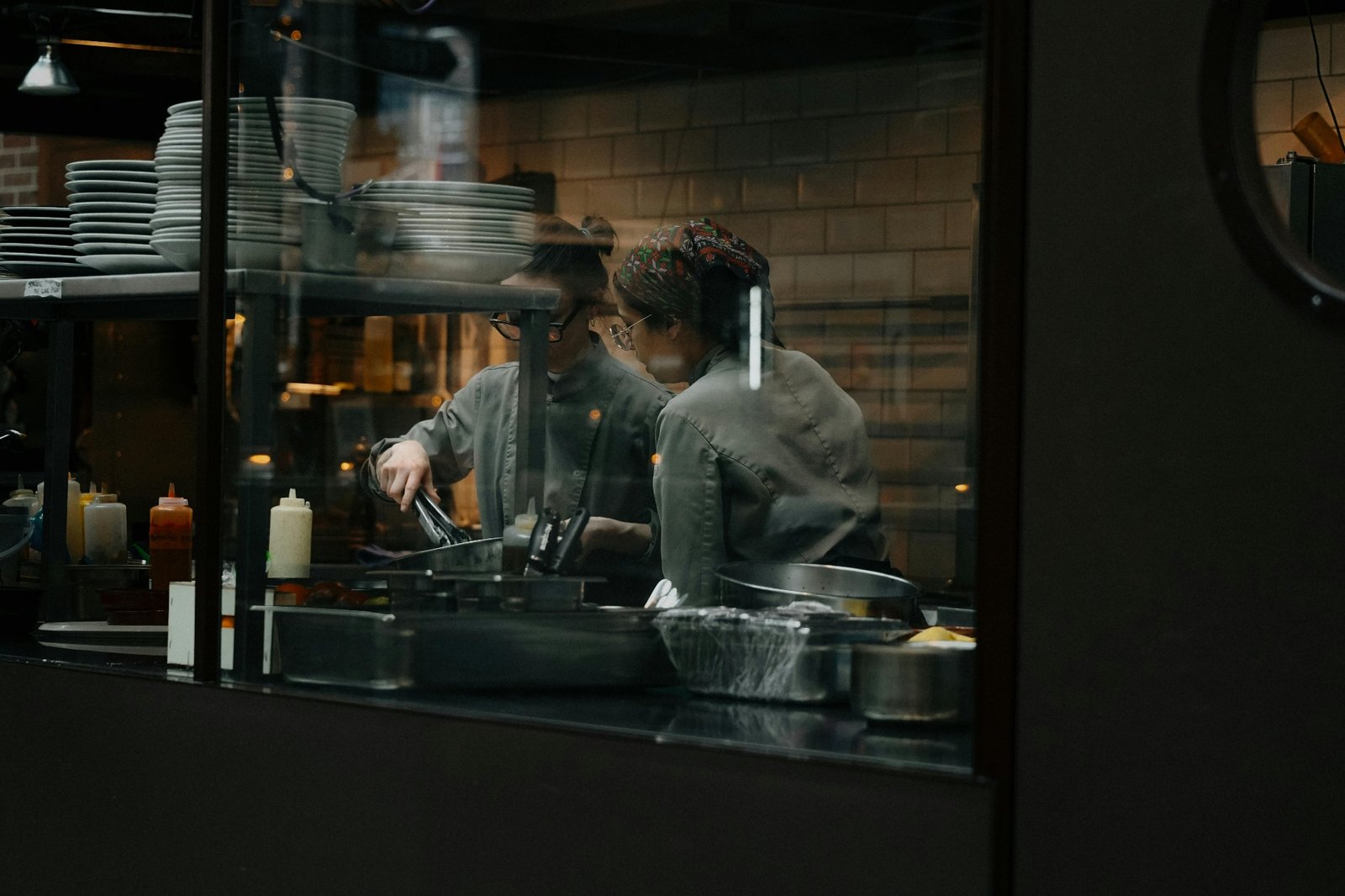 Two line cooks working back-to-back through a kitchen pass window during dinner service, juggling repetitive prep tasks that contribute to restaurant staff burnout