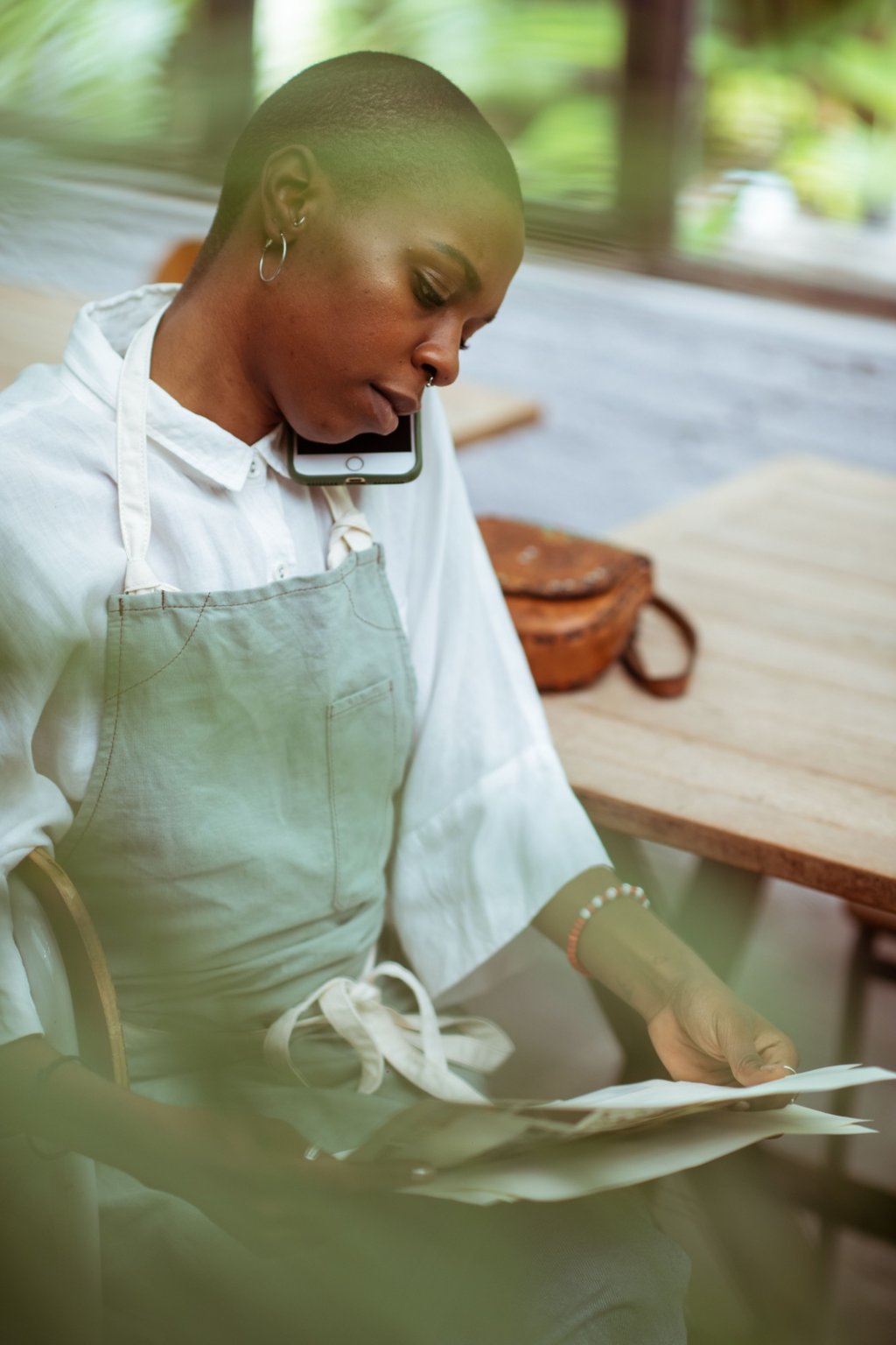 A restaurant owner in an apron on a phone call while reviewing paperwork at a table.