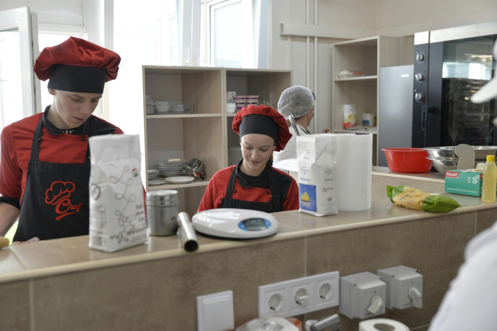 Two junior pastry cooks in chef hats training on prep, weighing ingredients and following recipe standards in a restaurant kitchen