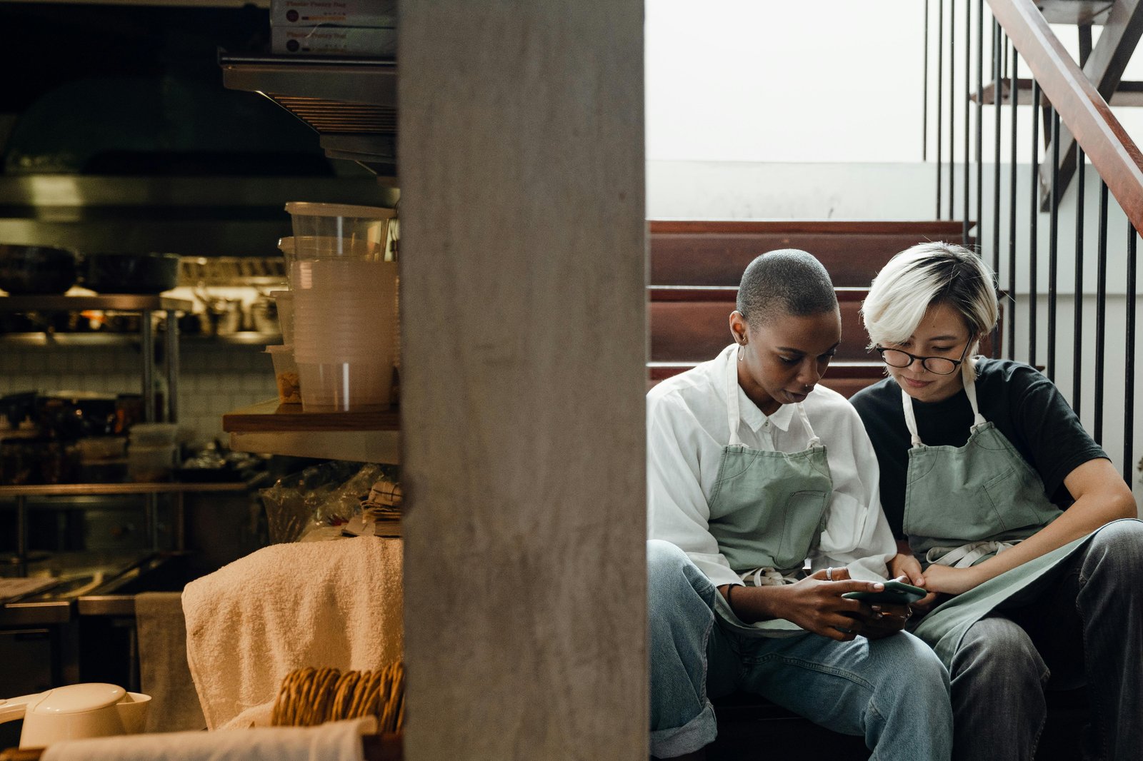 Two restaurant staff members in aprons taking a quick break together on the back stairs, checking a phone between shifts
