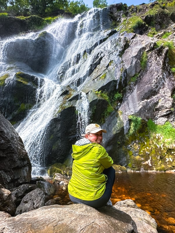 Women smiles towards the camera and sits at the bottom of a waterfall.
