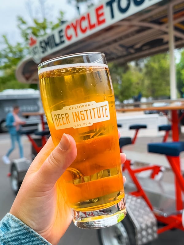 Woman holding a beer that says "Kelowna Beer Institute" with the Smile Cycle Tour bike in the background