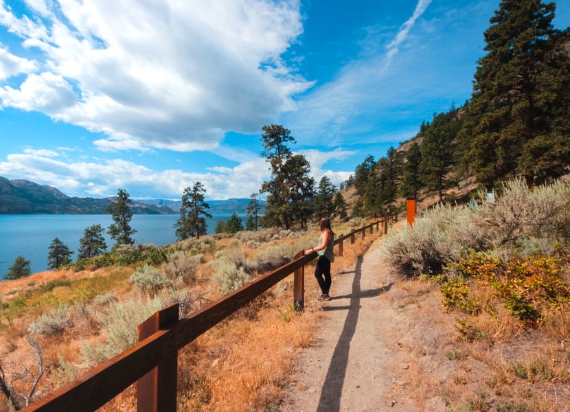 Woman on the Goat's Peak hiking trail, standing next to a wooden fence with views of Okanagan Lake.