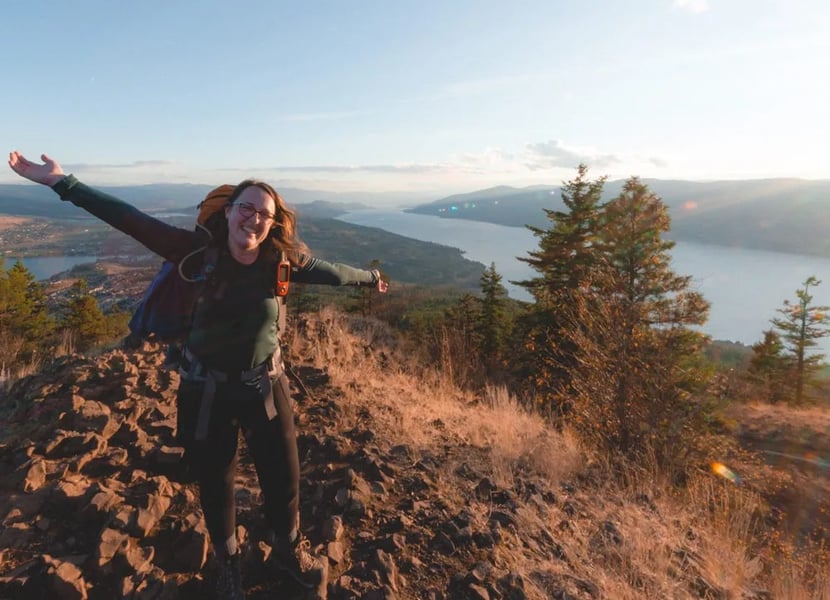 Woman carrying a large backpack stands with her hands wide open at the summit of Spion Kop at sunset