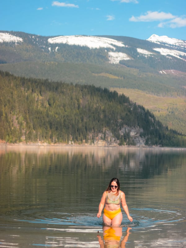 Woman in yellow bathing suit walks out of Arrow Lake with snow capped mountain in the background.