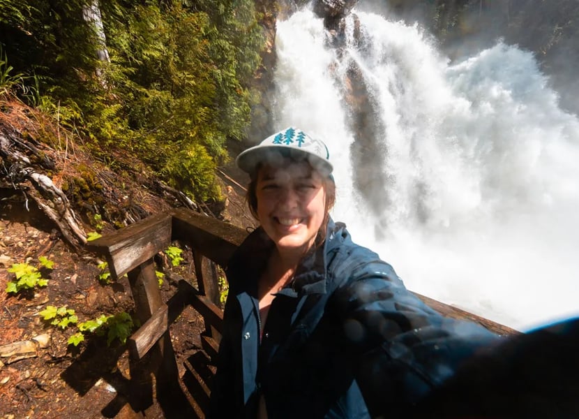 A woman wearing a hat and rain jacket takes a selfie in front of Rainbow Falls near Cherryville, BC.