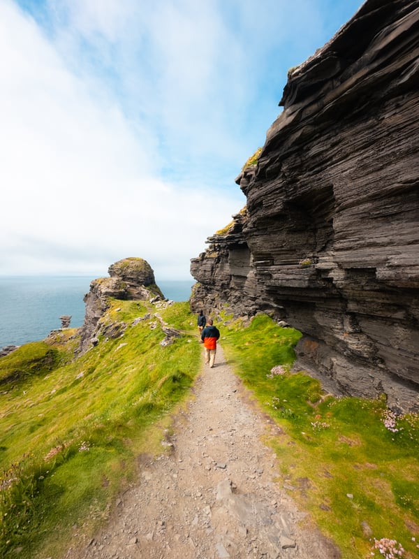 Two men walk on a narrow trail along a layered rock cliff at the Cliffs of Moher