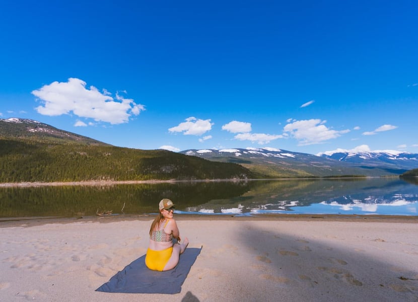 Woman in bathing suit sits on a towel on a sandy beach on Arrow Lake.