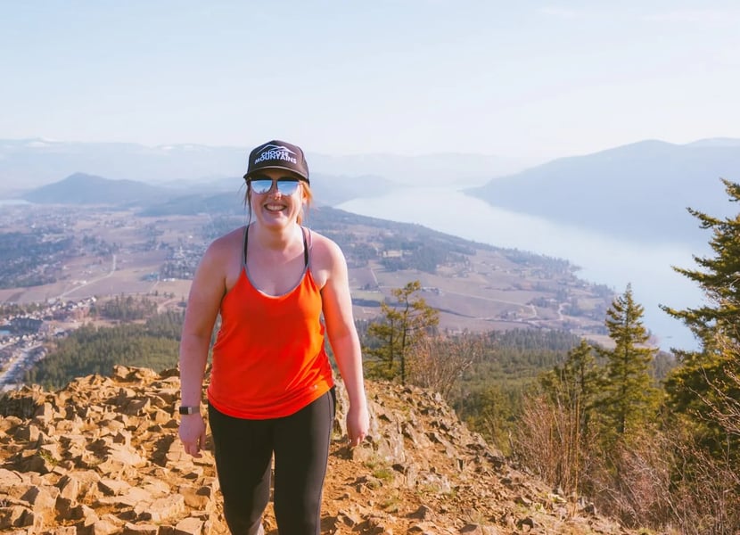 A woman in red walks towards the camera at the summit of Spion Kop. Lake Country, Kelowna, and Okanagna Lake are in the background.