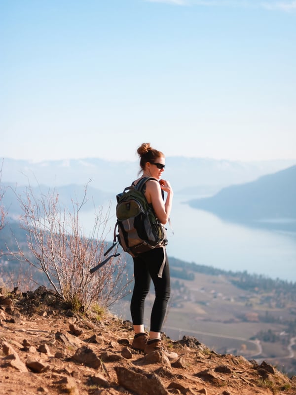 A women wearing a backpack and sunglasses looks out over Okanagan Lake from the summit of Spion Kop
