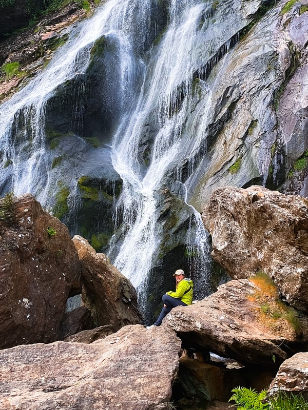 Woman sits at the base of a large waterfall.