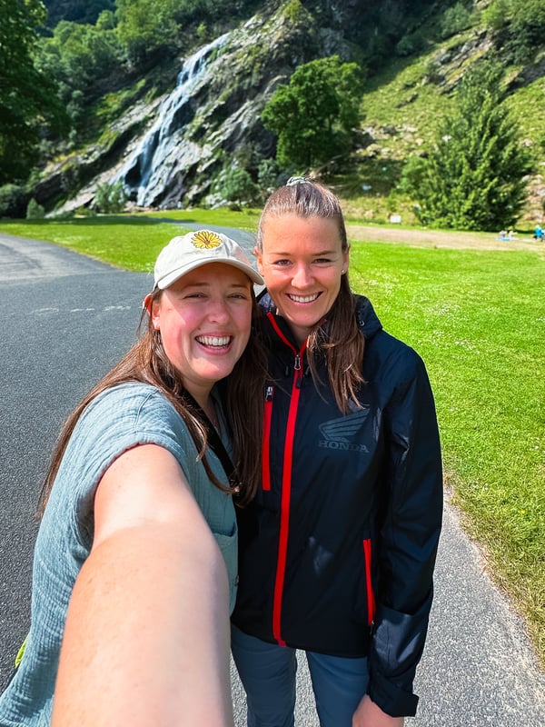 Selfie of two women standing on a paved trail in front of Powerscourt Waterfall.