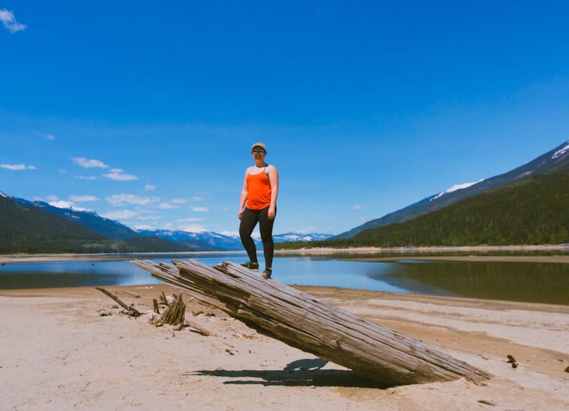 Woman in red tank top stands on top of a large piece of driftwood on the shores of Arrow Lake. Sunny day.