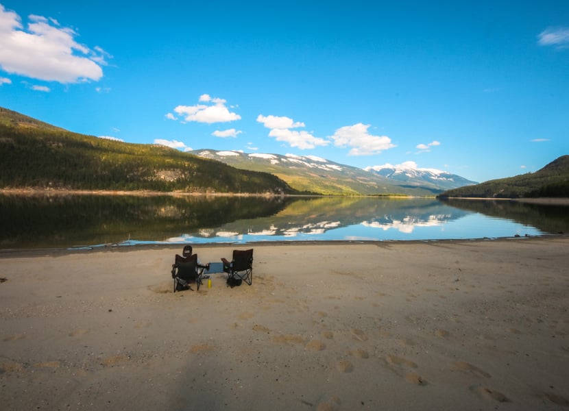 Man sits in one of two camp chairs on a sandy beach on Arrow Lake near Revelstoke BC. Sunny day with blue skies.