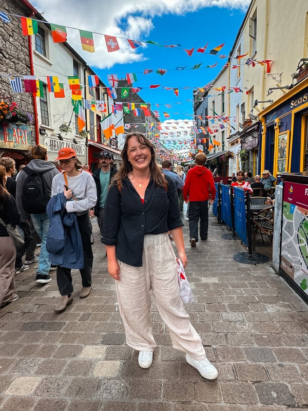A woman standing in a busy Irish walking street on a sunny day with flags in the background.