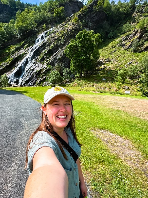 Selfie of a women in front of the waterfall while standing on a paved trail.